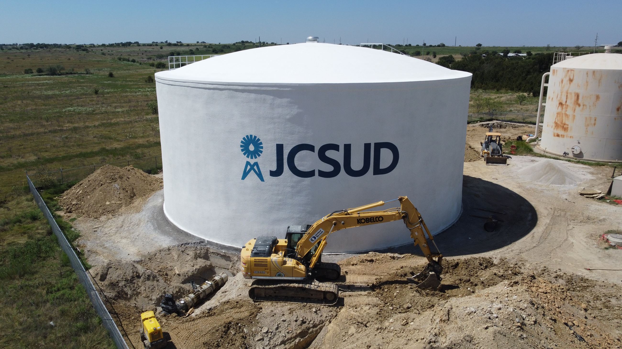 Image of excavator equipment infront of a white ground storage tank with the JCSUD logo